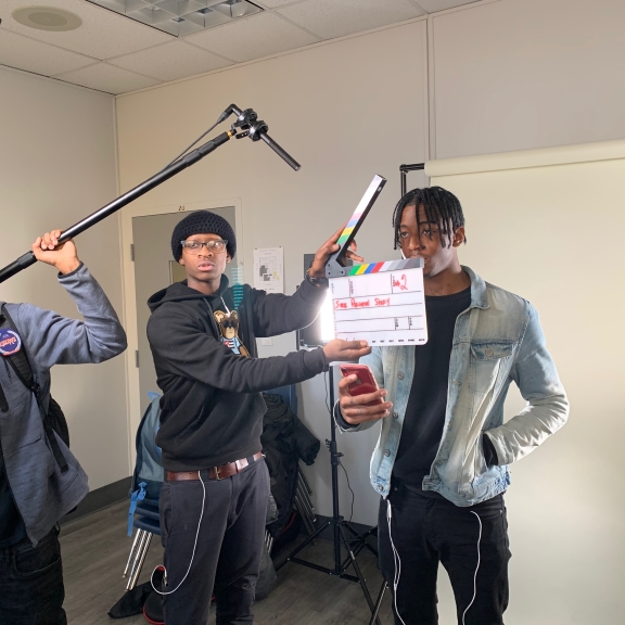 A person holds a boom mic while another uses a clapboard in front of someone about to be filmed