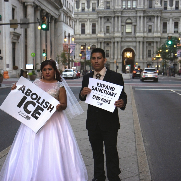 A bride holding a "Abolish ICE" sign and a groom with a "Expand Sanctuary. End PARS" sign, stand on a street partition in front of City Hall