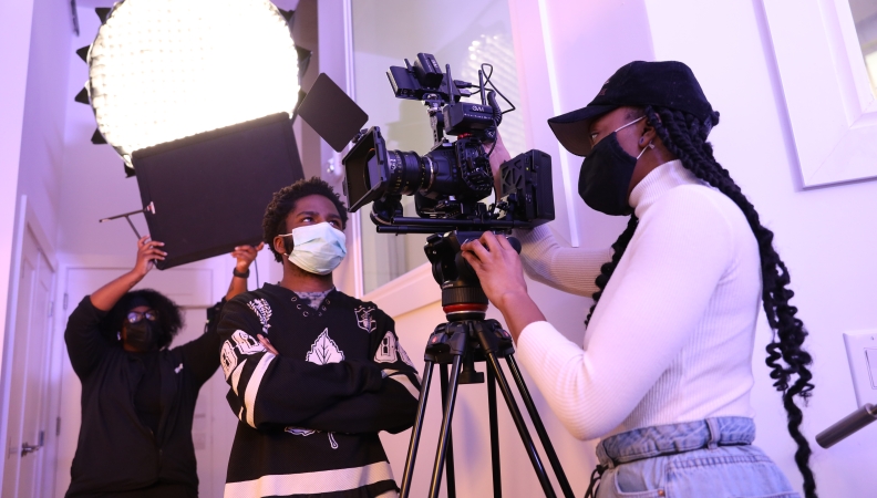 Two people talking around a video camera while another person holds up a black board in the background