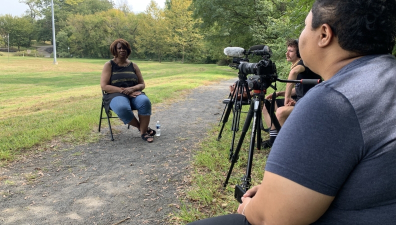 a woman sitting on a chair on a paved trail speaking with two seated people filming with cameras