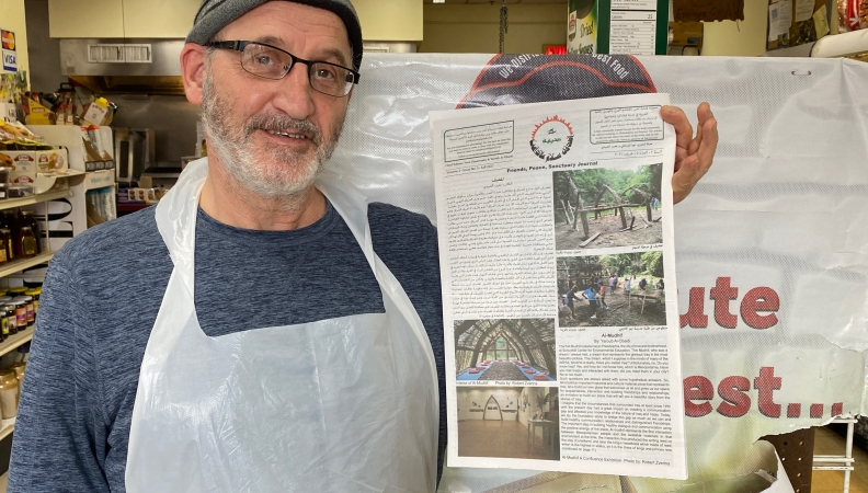 A close shot of a person wearing a gray shirt, gray knit hat and a plastic apron holds up a newspaper