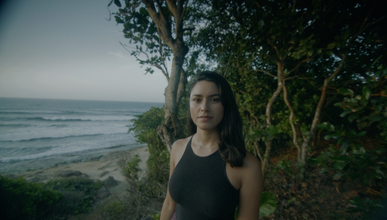 An olive toned person in a black halter top stands in front of trees on a beach