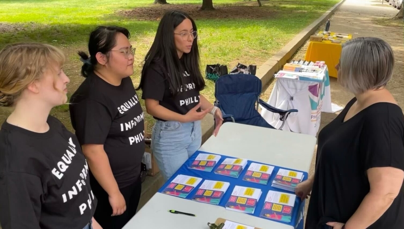 Three people wearing black t-shirts standing behind a table with printed cards as another person stands in front of the table