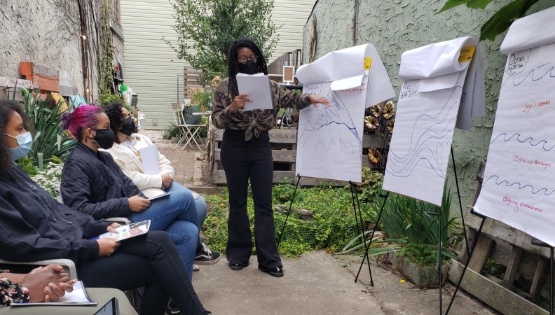 A person points to a drawing on a standing notepad in front of three sitting people