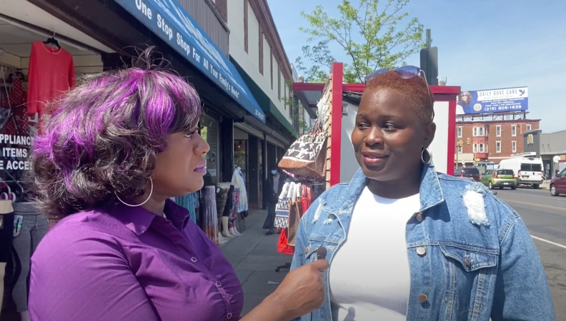 a woman with purple hair highlights and wearing a purple top interviews another woman wearing a jean jacket and a low cut fade hairstyle