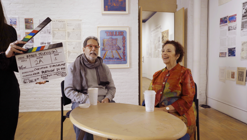 a behind the scenes film still of two people seated at a roundtable having a discussion and a person with a film slate standing to the left