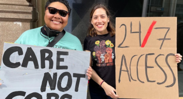 two people hold up signs that read "care not cops" and "24/7 access"