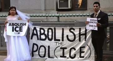 A bride and a groom each hold signs of protest against ICE while standing on opposite ends of a banner that reads "Abolish the Pol(ICE)"