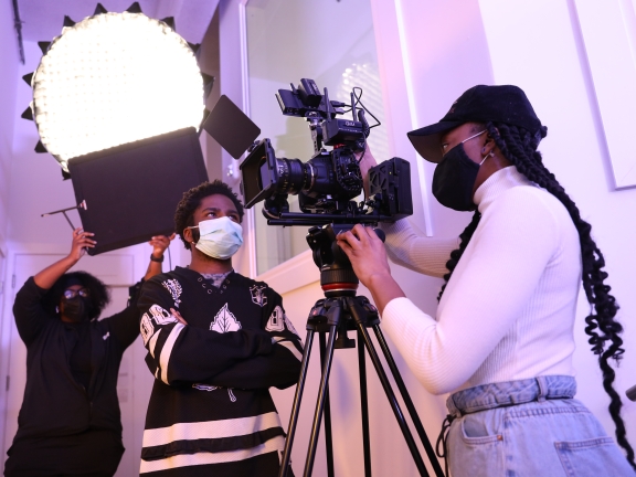 Two people talking around a video camera while another person holds up a black board in the background