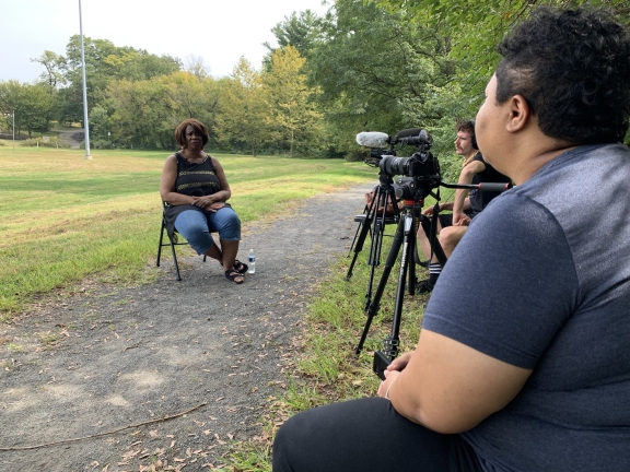 a woman sitting on a chair on a paved trail speaking with two seated people filming with cameras