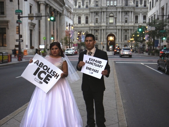 A bride holding a "Abolish ICE" sign and a groom with a "Expand Sanctuary. End PARS" sign, stand on a street partition in front of City Hall