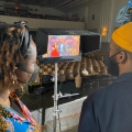 Two people looking at a monitor showing two people sitting in velvet chairs