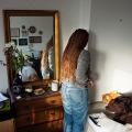 A person with brown braided hair, a gray top and blue jeans reaching into a corner past a dresser with a mirror on it