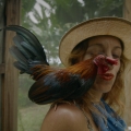 A person purses their lips towards a rooster perching on their shoulder