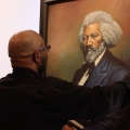 A bald person wearing glasses carrying a large portrait of Frederick Douglass in their hands