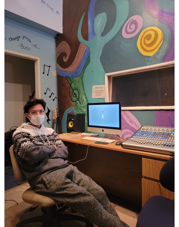 a masked person seated inside a music studio in front of a computer and audio equipment on a desk