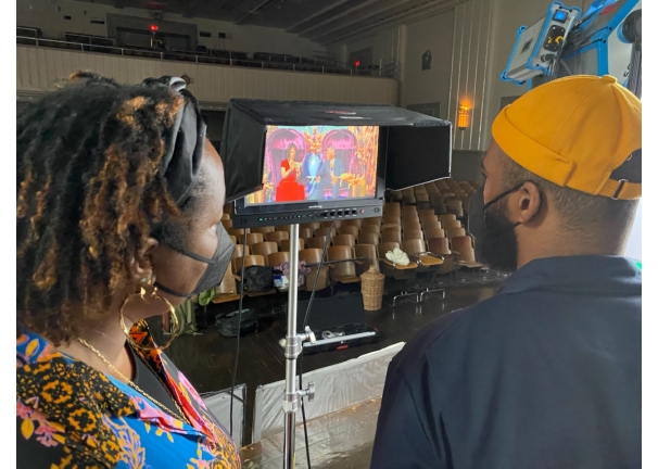 Two people looking at a monitor showing two people sitting in velvet chairs