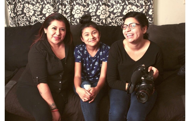 Three people, one of them a young person, sit on a cozy black sofa, smiling at the camera