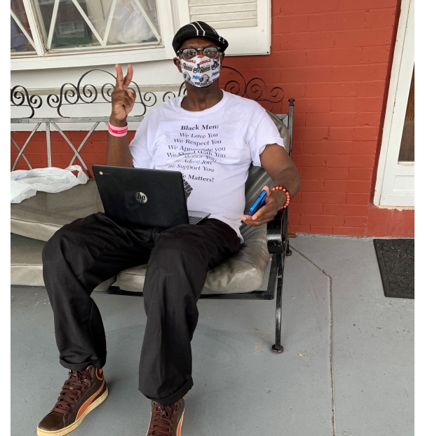 a man sitting on an outdoor couch with a laptop in his lap and phone in his hand giving a peace sign