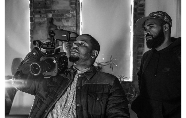 Three people looking at the viewfinder of a video camera one person has on their right shoulder