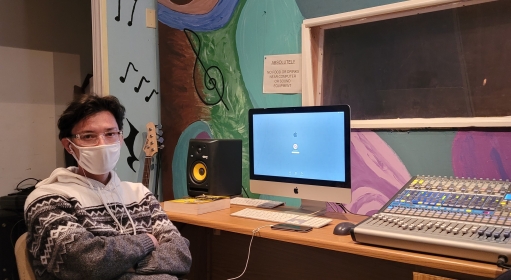 a masked person seated inside a music studio in front of a computer and audio equipment on a desk