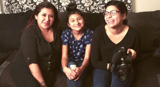 Three people, one of them a young person, sit on a cozy black sofa, smiling at the camera