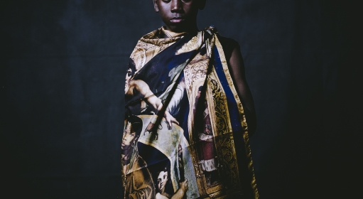 A dark toned child wearing a gold and blue large scarf with religious figures tied around their body