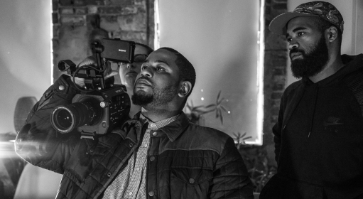 Three people looking at the viewfinder of a video camera one person has on their right shoulder