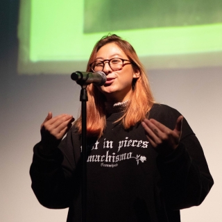 woman wearing glasses stands as she speaks into a microphone